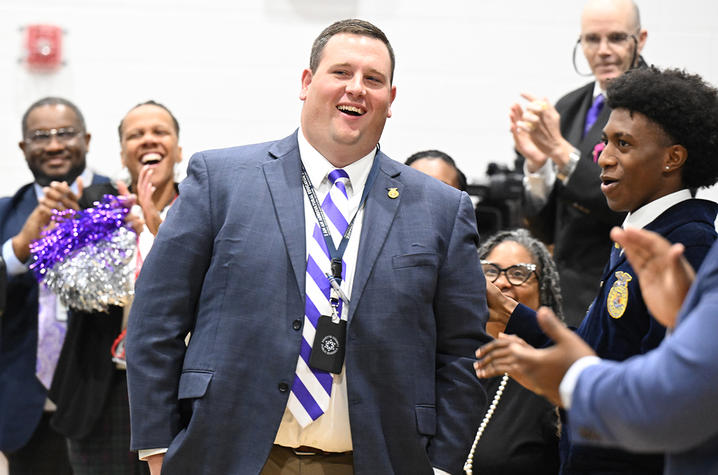 man in suit with white and purple tie standing in center of crowd that is cheering for him
