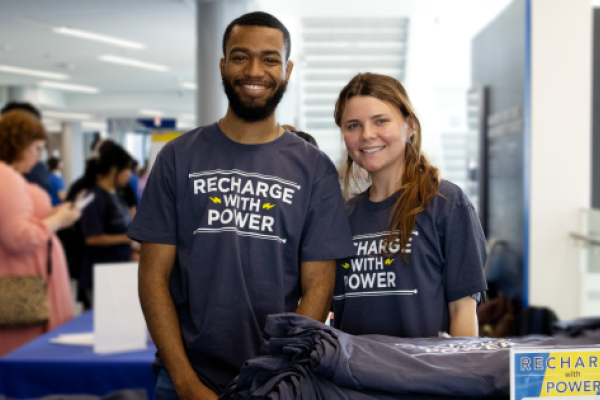 two students standing next to each other in the student center with shirts that say "recharge with power"