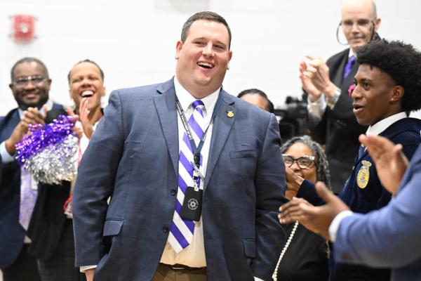 man in suit with white and purple tie standing in center of crowd that is cheering for him