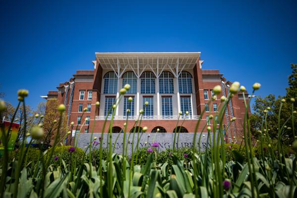 a low-angle view of the front of William T. Young library with flowers and grass in the foreground and a clear blue sky